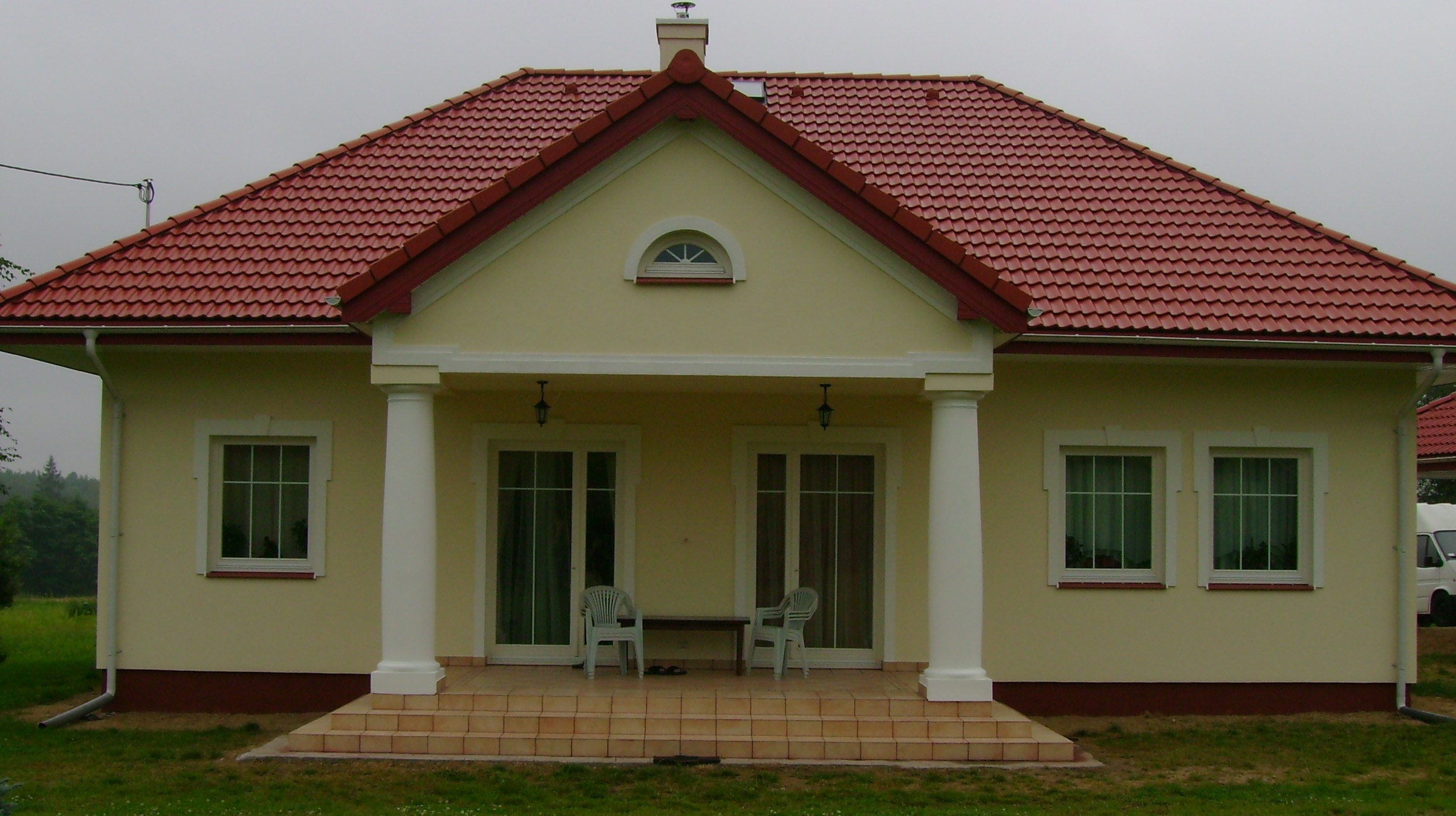 Jasnożółty dom jednorodzinny z czerwoną dachówką, białymi kolumnami przy wejściu i dwoma plastikowymi krzesłami na tarasie.